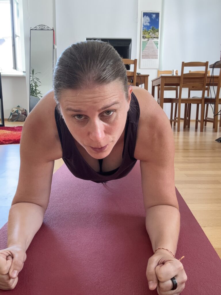Generose in Plank-Haltung auf ihrer roten Yoga-Matte. Im Hintergrund seiht man das Zimmer mit Tisch, Stühlen, Spiegel und Bild an der Wand.