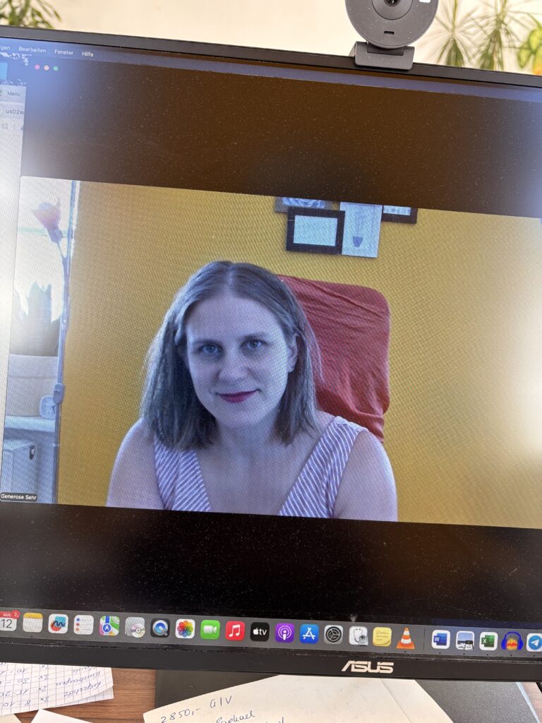 Generose hat gerade ein Zoom-Meeting gestartet und hat sich selbst über den Bildschirm fotografiert. Im Hintergrund eine gelbe Wand mit einem Bilderrahmen, ein Stuhl mit einem orangenen Überzug.