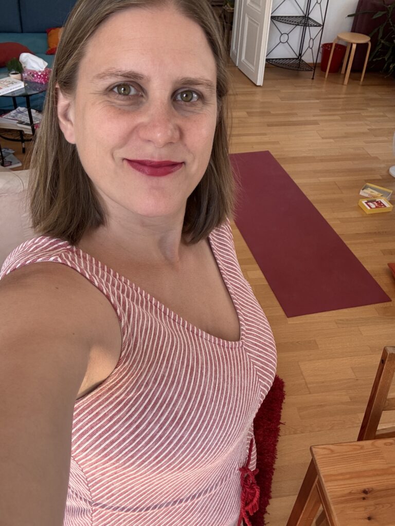 Ein Selfie von Generose. Sie hält die Kamera oben und schaut rein. Sie trägt ein rot-weiß gestreiftes Kleid mit V-Ausschnitt und roten Lippenstift. Im Hintergrund ist die Yogamatte und sonstige Teile des Zimmers zu sehen (Tisch, Regal etc.)