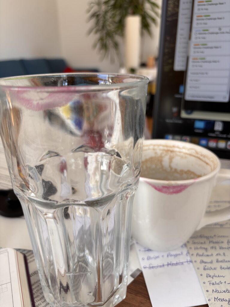 Ein Glas und eine Kaffeetasse, beides mit Lippenstift-Spuren. Auf der Seite der Bildschirm vom Computer und im Hintergrund die Pflanzen im Zimmer und ein Sofa.