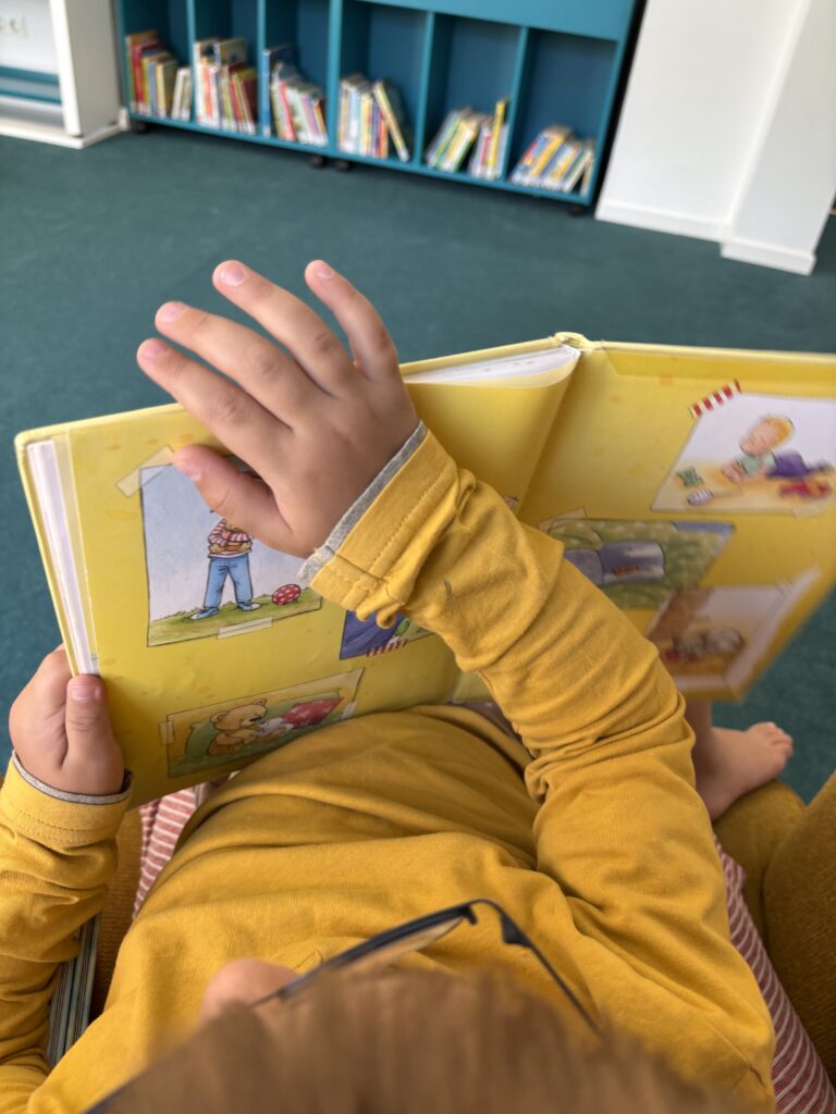 Auf dem Bild sieht man ein kleines Kind von oben fotografiert, das ein Buch anschaut und gerade umblättert. Das Kind trägt ein gelbes Shirt. Im Hintergrund sind Bücherregale und ein petrolfarbener Boden zu sehen.
