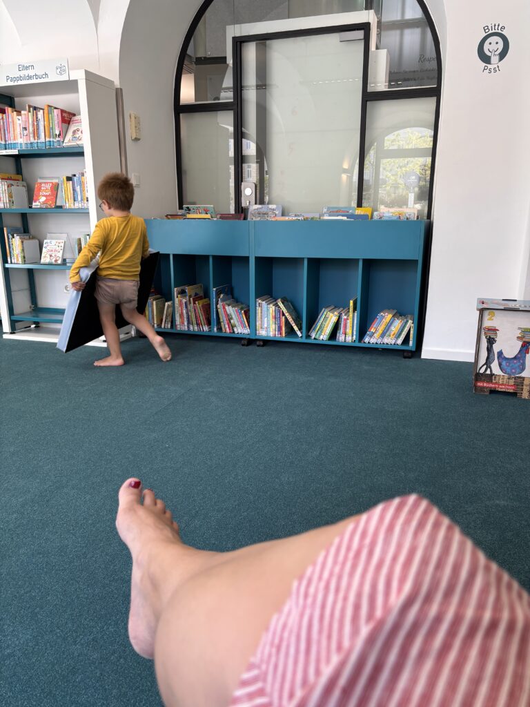 Ein weiteres Foto aus der Bücherei. Dieses Mal sieht man Generoses überschlagene Beine im rot-weiß gestreiften Kleid. Und im Hintergrund das Kind beim Bücherregal.