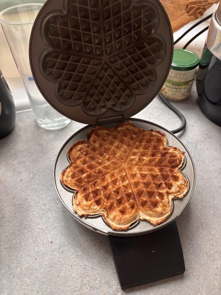 Ein geöffnetes Herz-Waffeleisen mit einer fertig gebackenen Waffel. Auf einer grauen Arbeitsplatte. Im Hintergrund sieht man diverse Gläser.