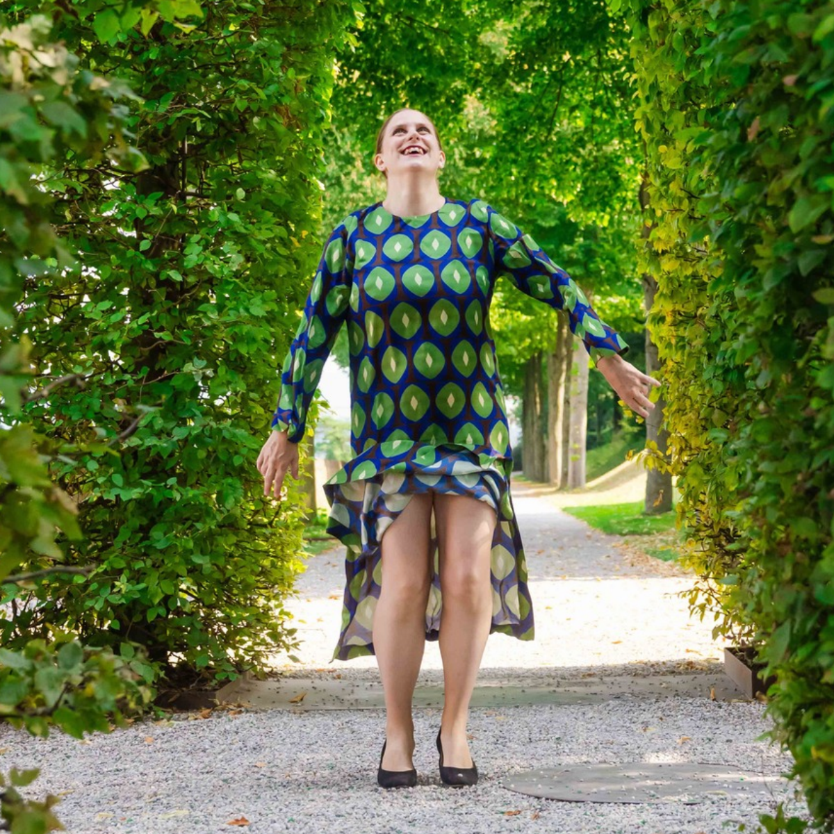 Generose steht singend da die Hände vor der Brust offen ineinander gelegt. Die Augen geschlossen. Sie trägt ein gepunktetes Kurzarm-Oberteil und einen lila Grütel. Im Hintergrund sind zwei Orgelpfeifen zu sehen. 